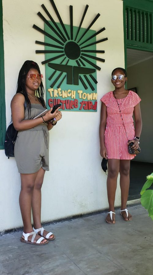 Young ladies standing in front of Trench Town Culture Yard Sign, Kingston, Jamaica